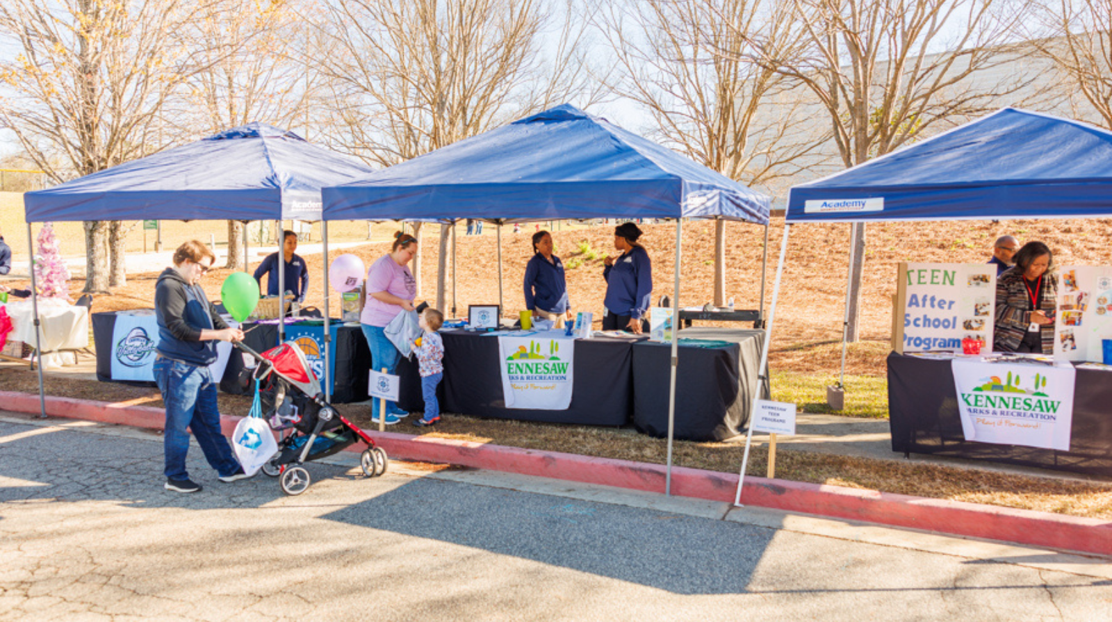 A group of people gathers around a table with a baby stroller, surrounded by multiple tables and tents in the background.