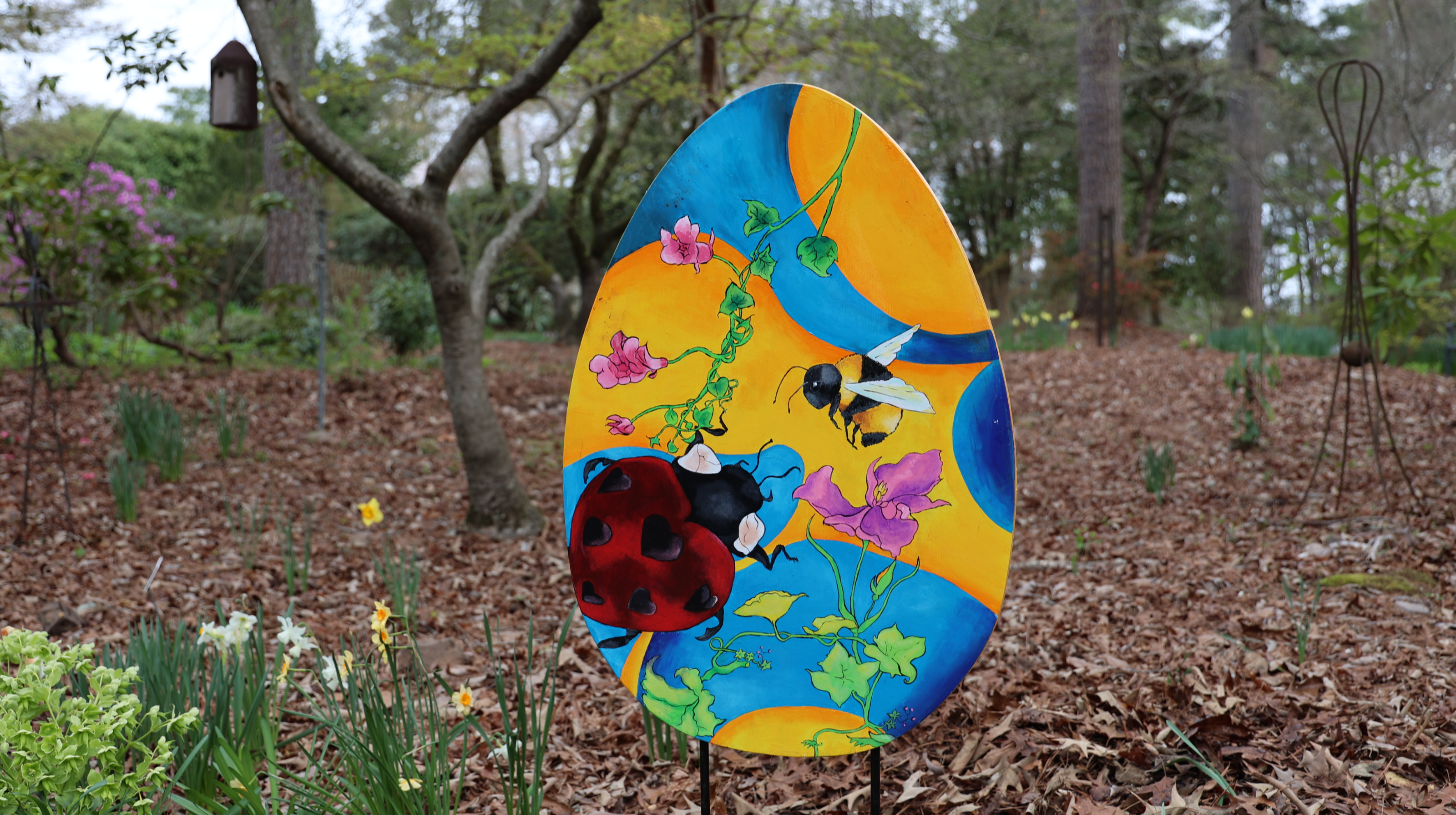 A brightly colored painted egg stands in a garden, showcasing illustrations of ladybugs, bees, and pink flowers against a blue and yellow background. The garden is filled with green foliage and leaves on the ground, creating a natural setting.