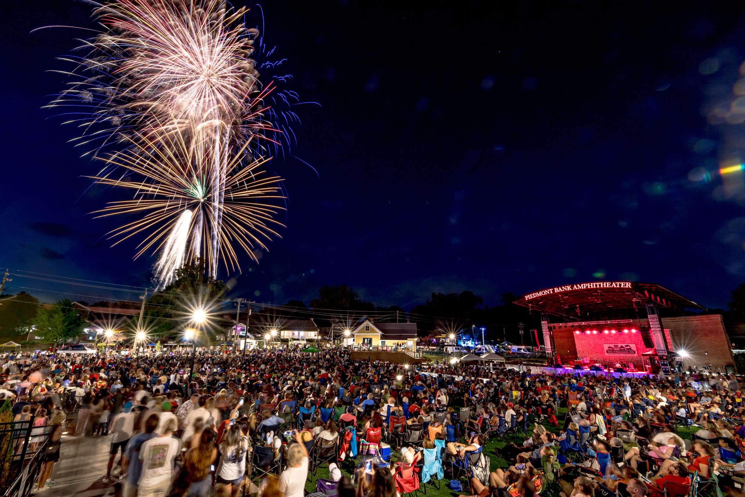 A large crowd gathers at night, gazing up at vibrant fireworks lighting up the sky.