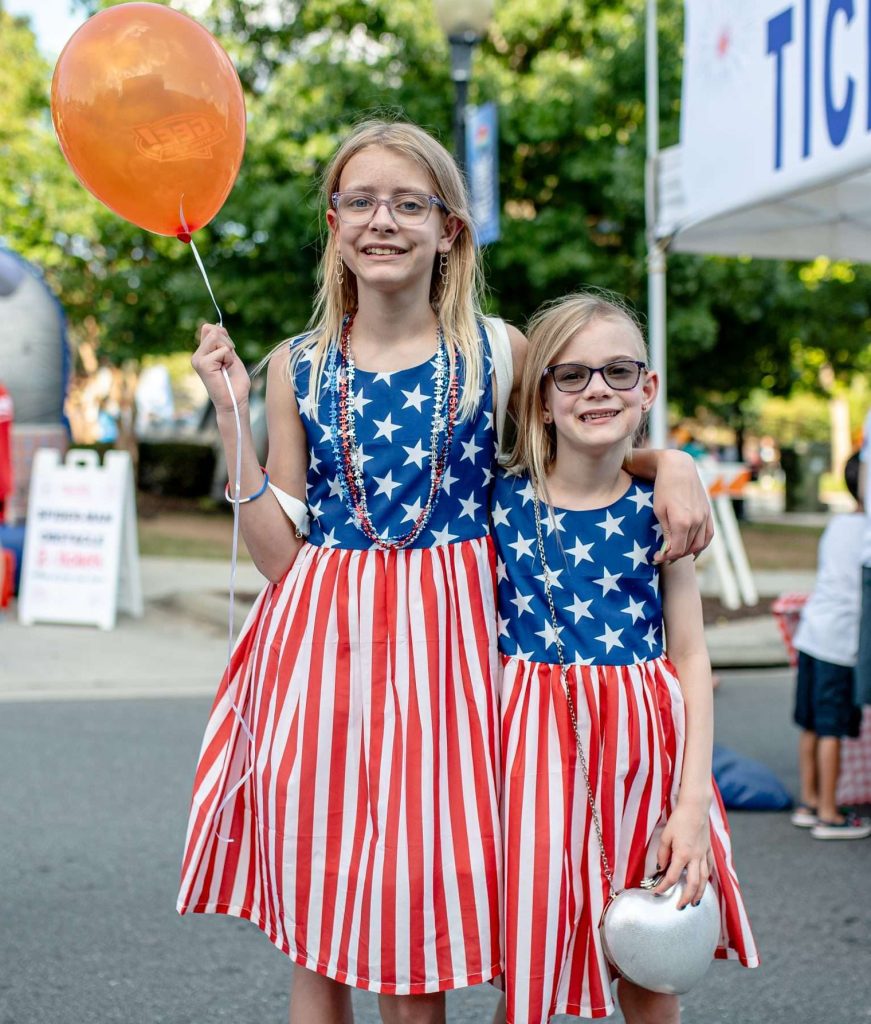 Two girls wearing patriotic dresses smile and wave at a parade, surrounded by colorful decorations and festive atmosphere.