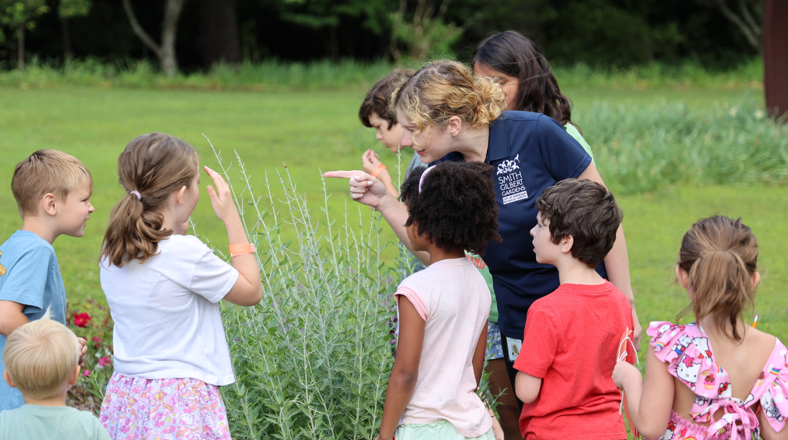 A woman pointing at a a ladybug on a plant and showing it to a group of kids in a garden