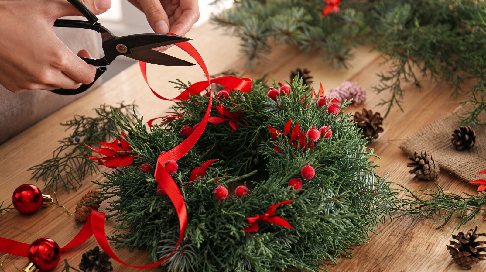 A holiday wreath laying on a table with someone's hands holding a red ribbon and scissors