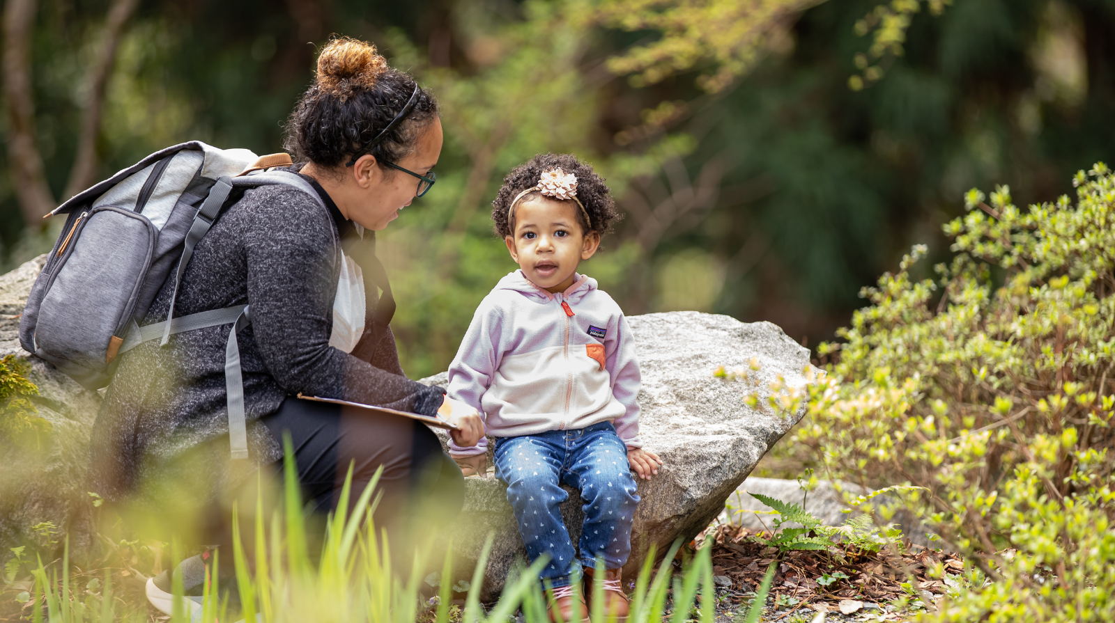 A women smiling down at a young girl sitting on a large rock in a garden