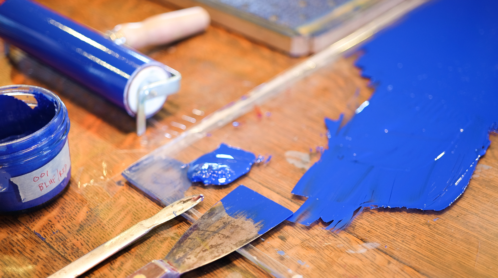 A wooden table with vibrant blue ink spread out using a spatula, next to a container and roller. The scene conveys creativity and artistic focus.
