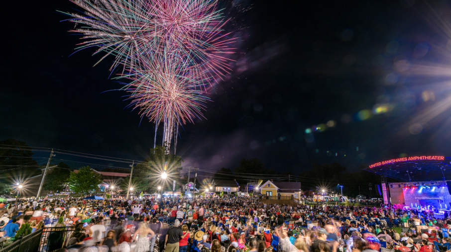 A vibrant fireworks display lights up the night sky over a crowded outdoor amphitheater. The energetic crowd watches in awe, creating a festive atmosphere.