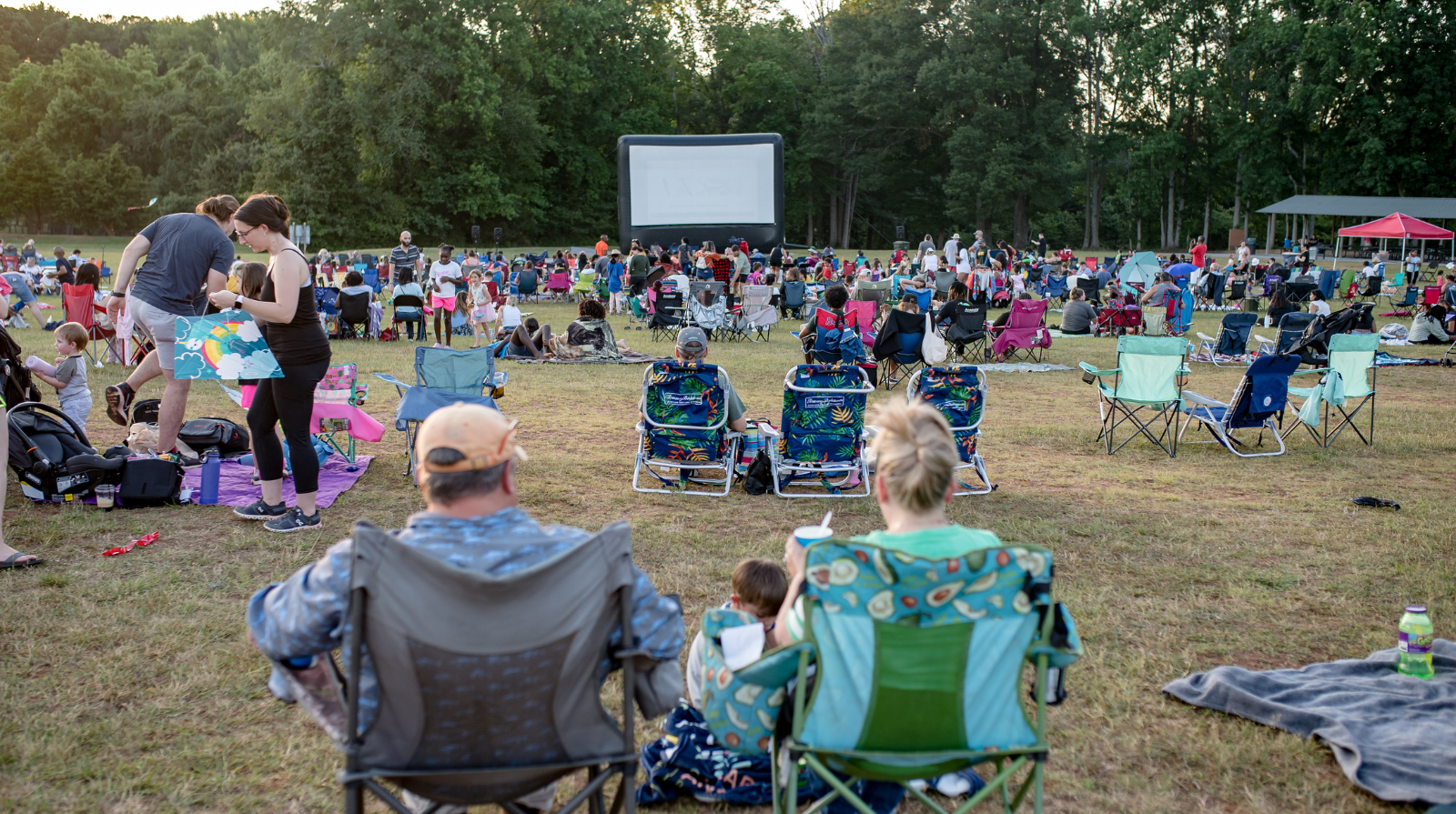Outdoor movie night scene with a large inflatable screen and a crowd lounging on foldable chairs on a grassy field, surrounded by trees at dusk.
