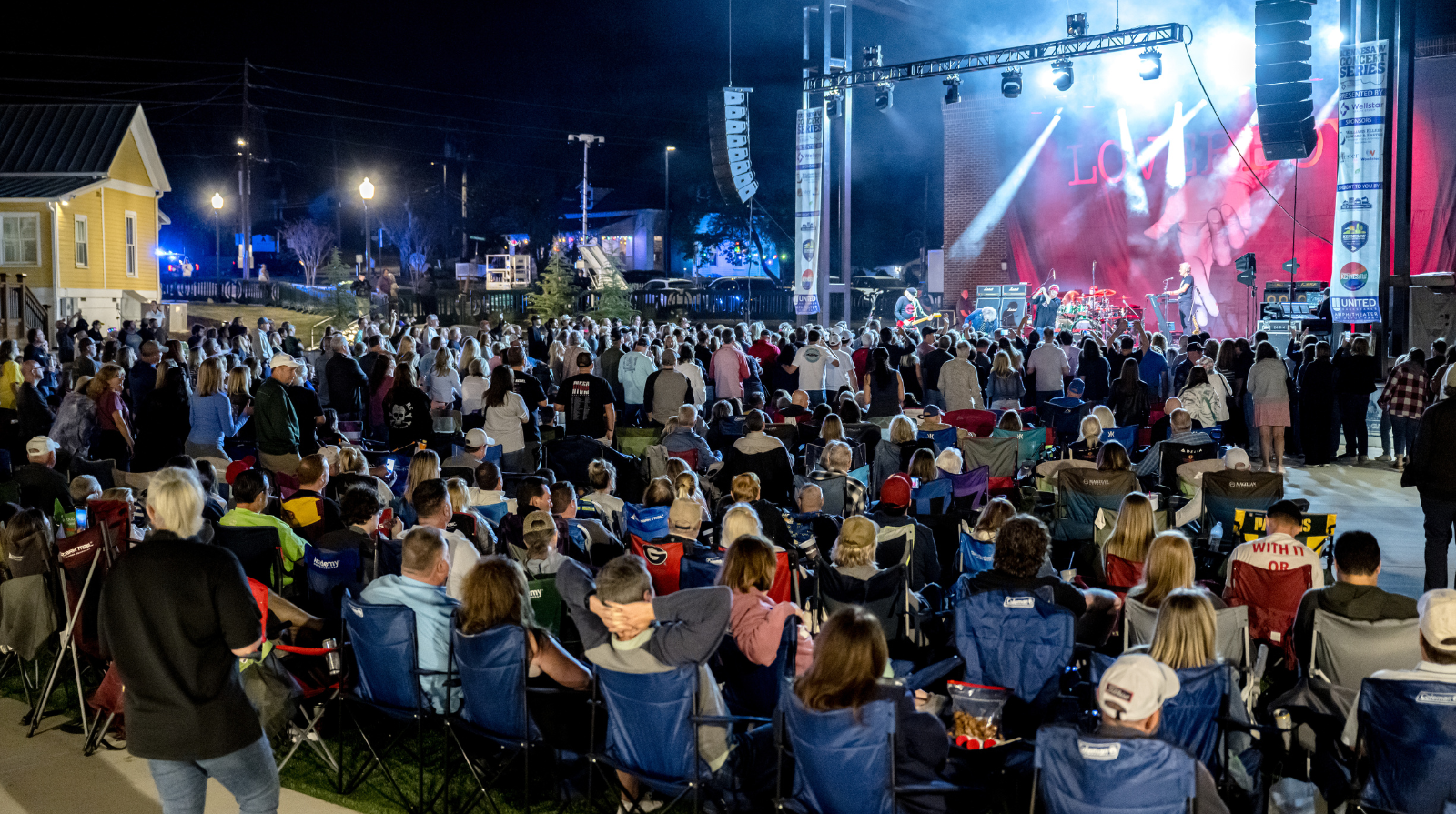 A large crowd of people enjoying an outdoor concert, with colorful lights and a stage in the background.