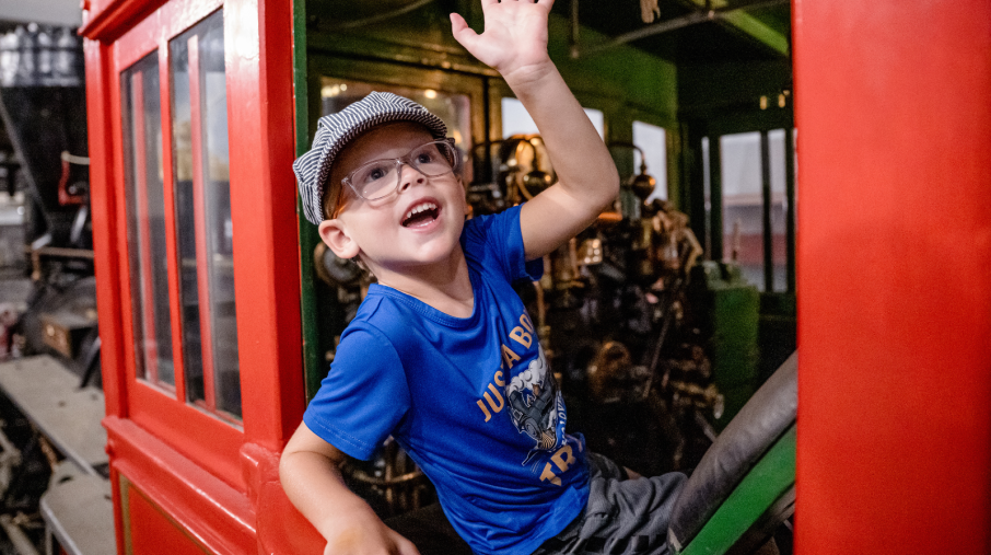 A joyful young boy wearing glasses and a cap waves enthusiastically from inside a vibrant red and green train cab. The scene conveys excitement and playfulness.
