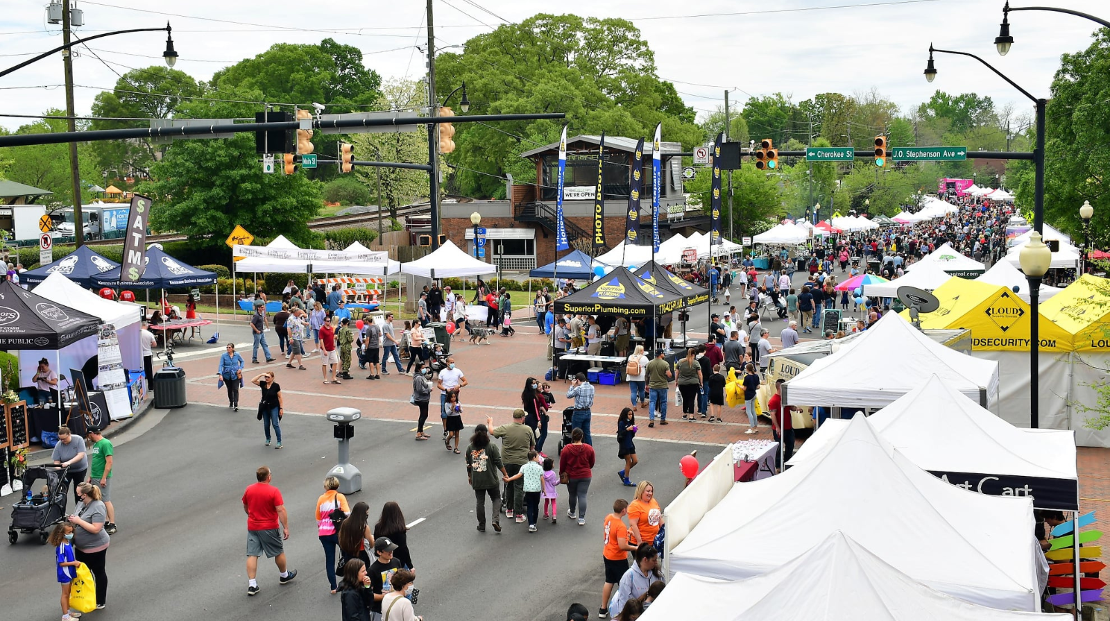 A street festival with rows of vendor tents, people walking and gathering, trees in the background, and traffic signals overhead.