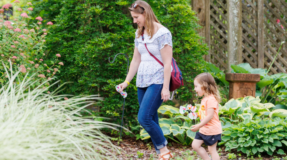 A woman and a child walk through a lush garden, each holding colorful pinwheels. The scene is joyful and vibrant, with greenery and blooming flowers.