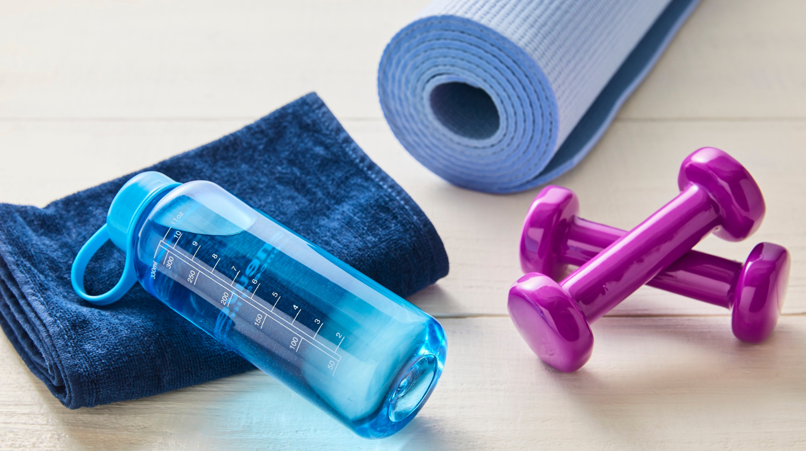 Fitness setup with a rolled blue yoga mat, purple dumbbells, a blue towel, and a translucent blue water bottle on a light wooden floor.
