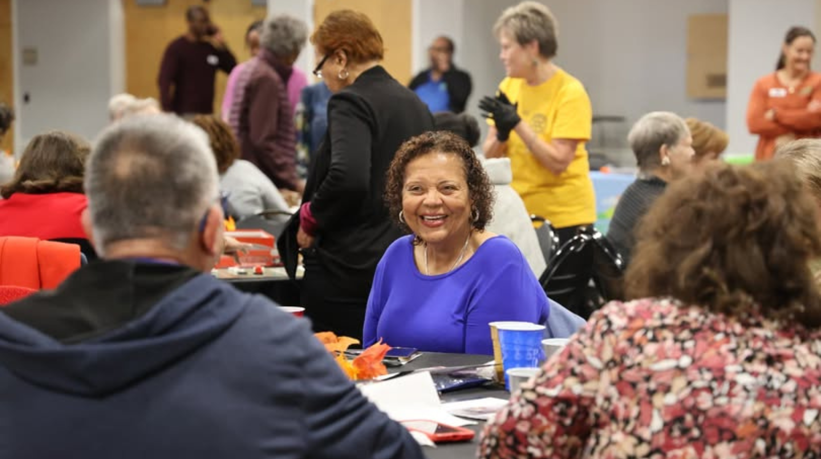 A woman in a blue shirt smiles warmly at a lively gathering. People are seated at a table, engaged in conversation, creating a cheerful atmosphere.