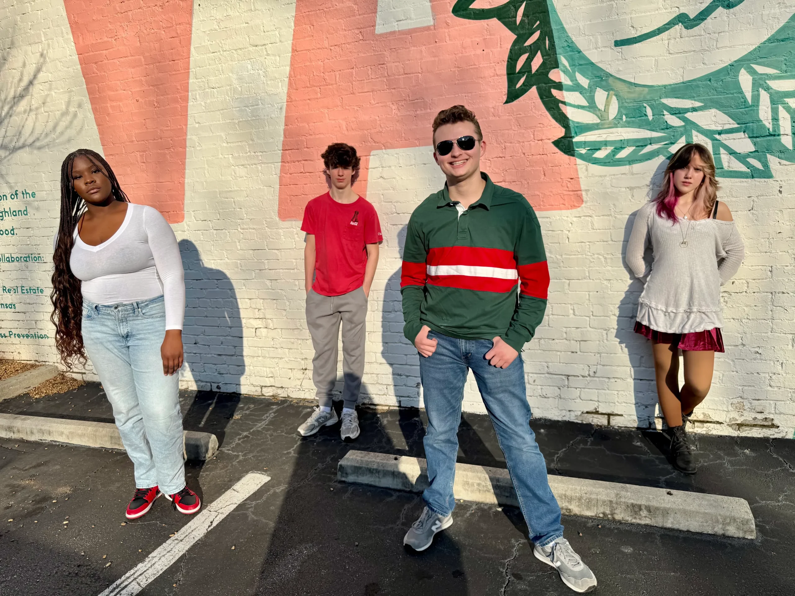 Four young adults stand casually against a brick wall mural. The sun casts strong shadows, creating a relaxed and vibrant atmosphere.