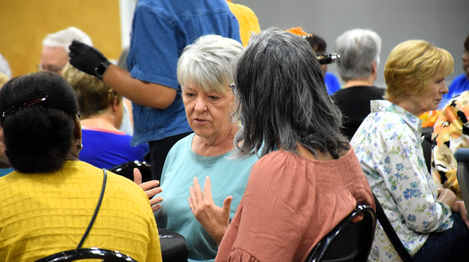 Two women sit and talk in a crowded room, surrounded by other people engaged in conversation.