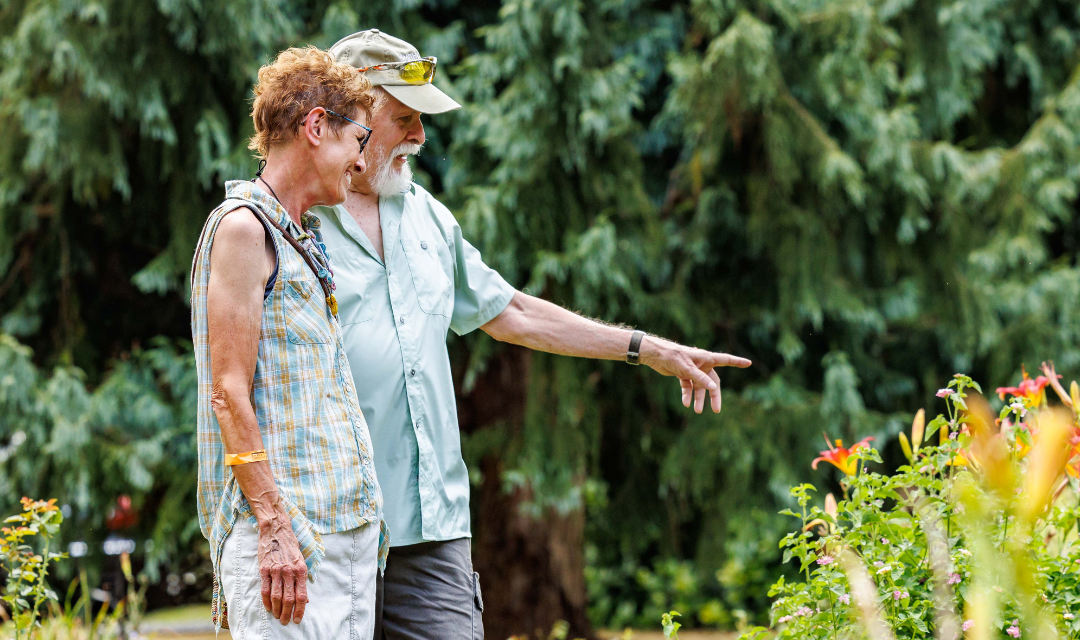 A man is pointing at flowers in a garden while talking to a woman, both smiling and engaged in conversation.