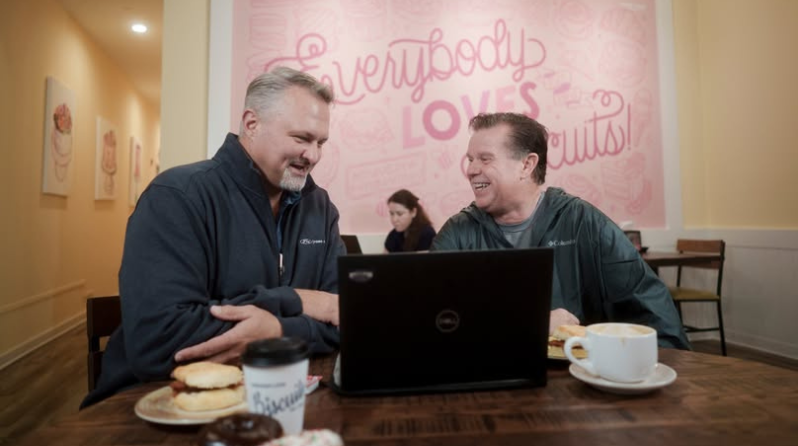 Two men smiling and discussing over a laptop in a café, with pastries and coffee on the table, and a light pink mural reading “Everybody loves donuts!” in the background.